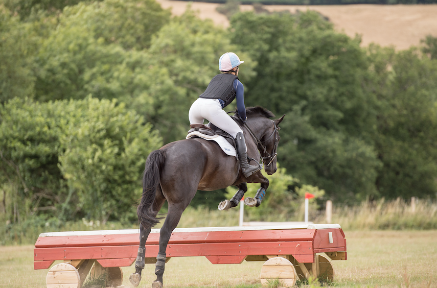 A rider is pictured wearing a black body protector while jumping a cross-country jump on a dark horse