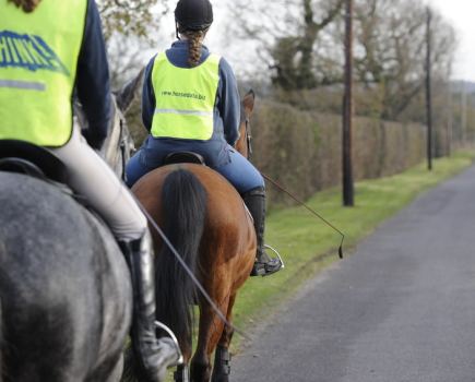 The images shows two riders on the roads. Both are wearing high vis; one is riding a grey horse, the other is riding a bay horse.
