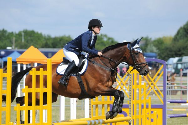 Pictured is a rider on a bay horse and rider clearing a yellow showjumping while competing on grass